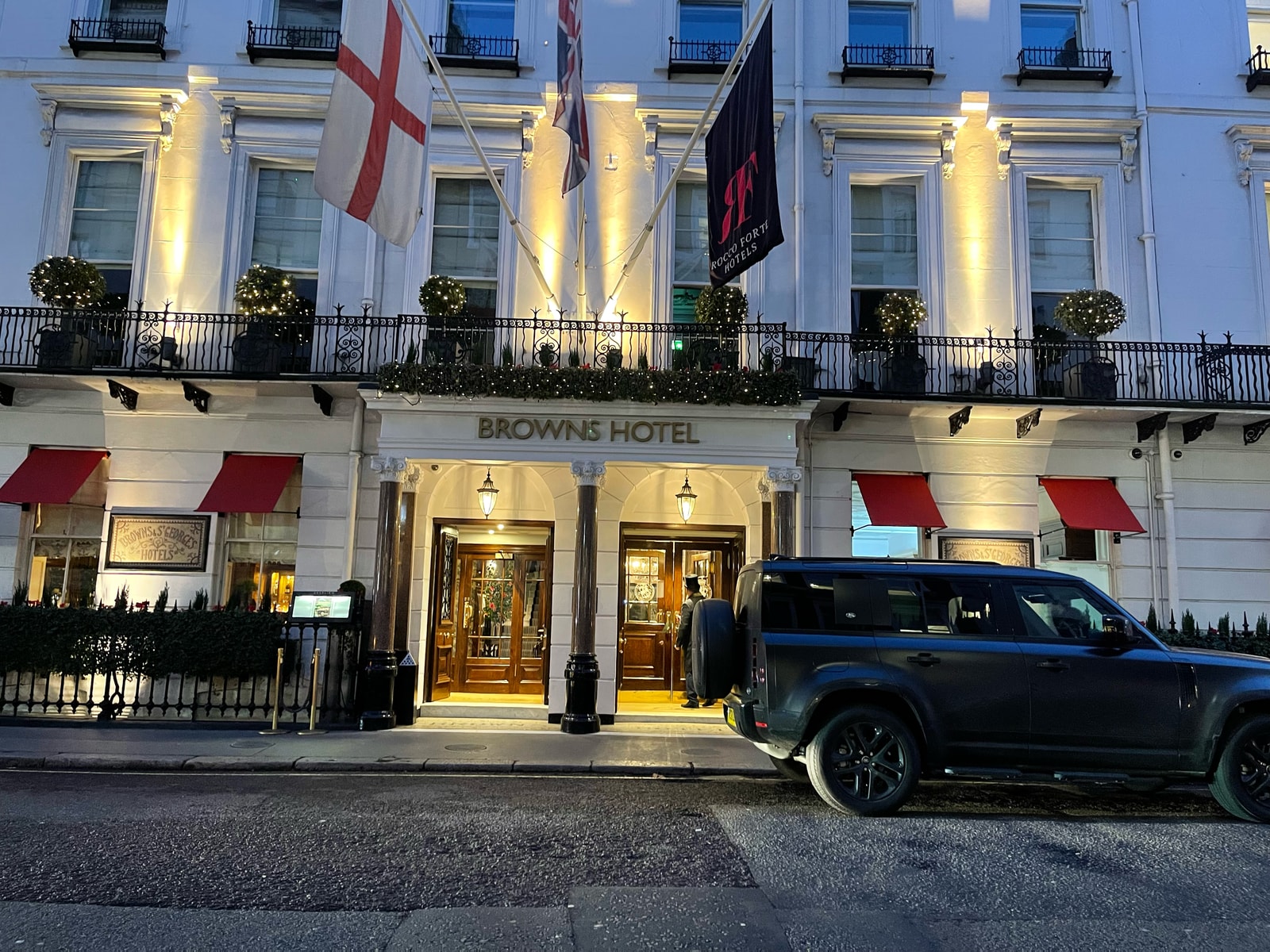 Brown's Hotel in Mayfair at dusk, its entrance illuminated beneath the flags. Our photograph of the hotel exterior.