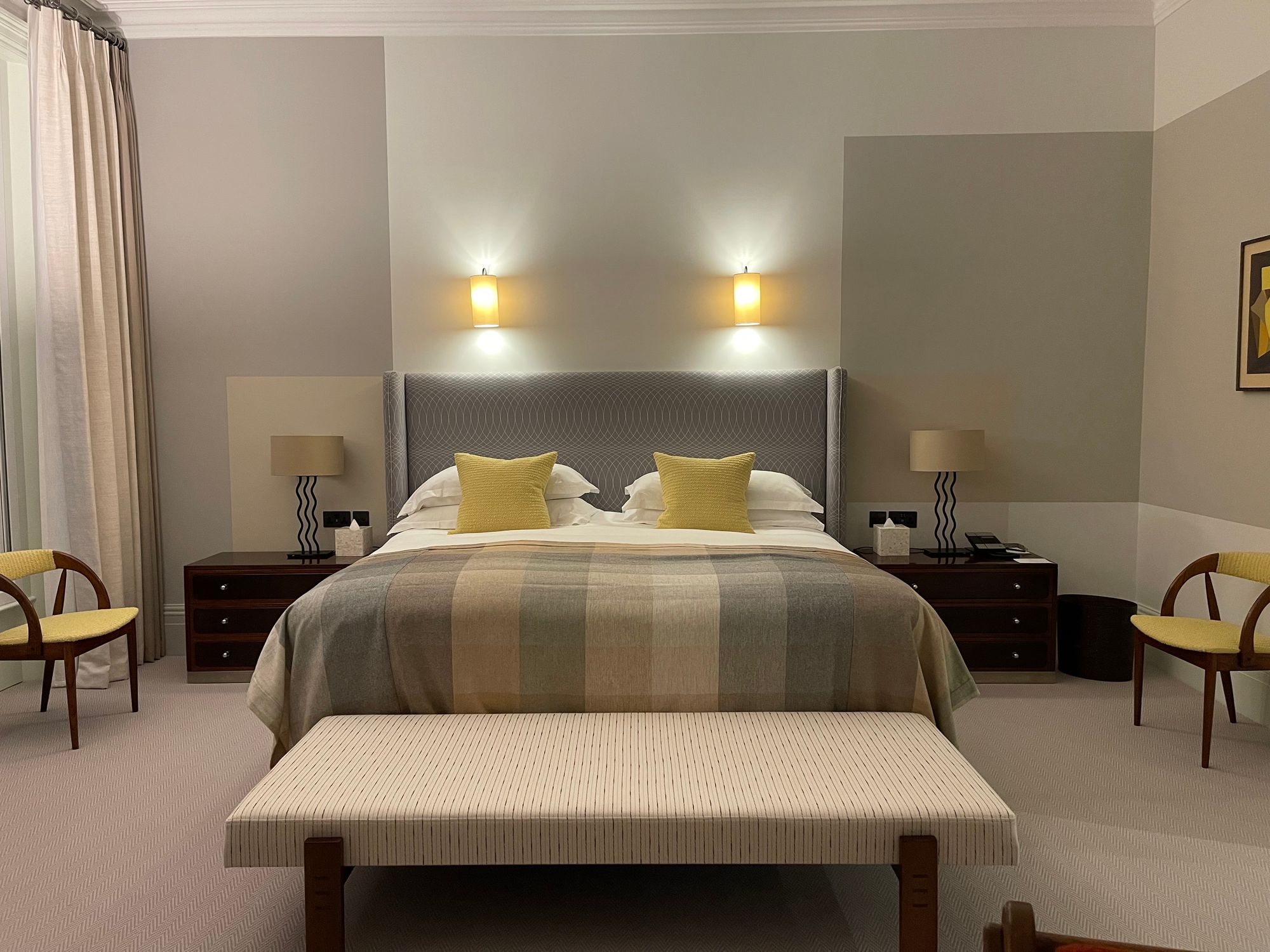 Finished guest bedroom at Brown's Hotel, Mayfair: grey upholstered headboard, yellow cushions, a checked throw and twin wall lights on pale grey painted walls.