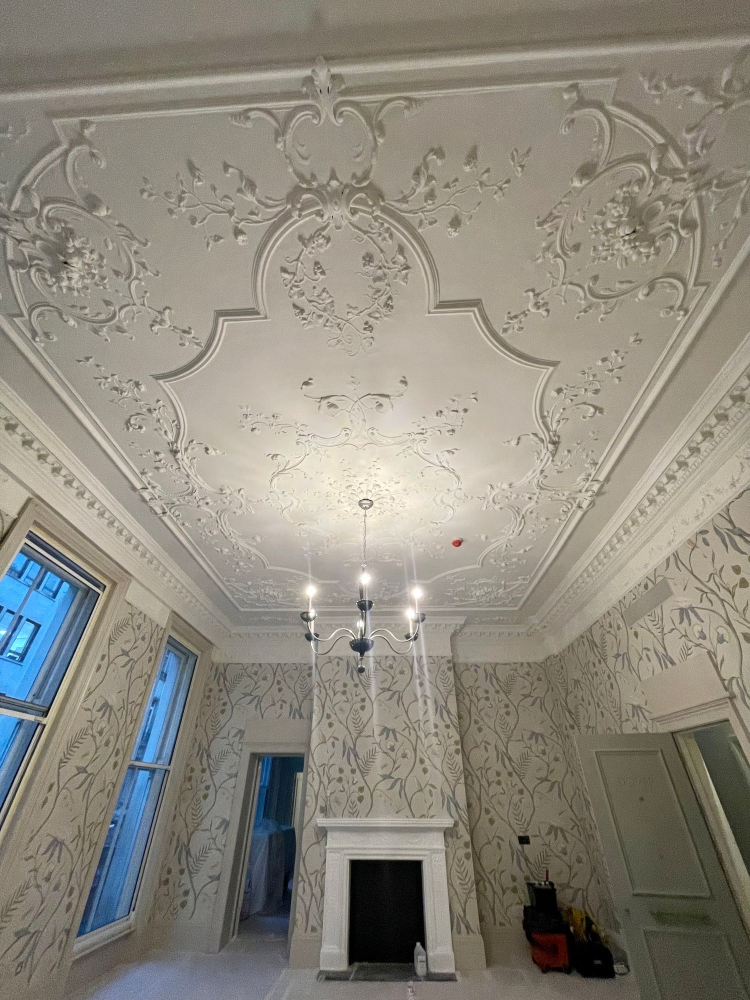 Ornate plaster ceiling and chandelier above walls hung in Adam's Eden wallcovering at Brown's Hotel, Mayfair, photographed during works with floor protection and equipment by the door.