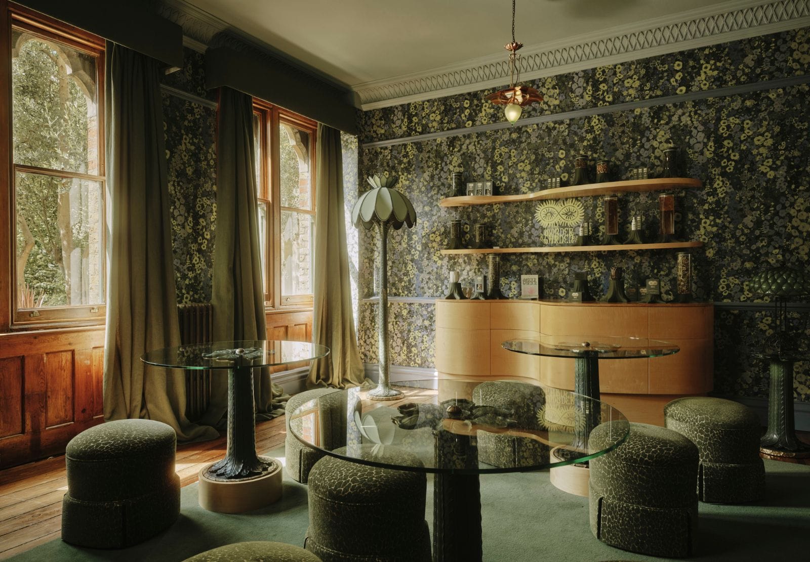 Green-and-gold floral wallpaper above timber panelling in a finished room at the House of Hackney showroom, with glass tables and low stools.