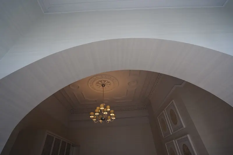 A wallcovered arch seen from below, its curved outer edge finished as one crisp, even line, framing a room with a decorative plaster ceiling and a chandelier.