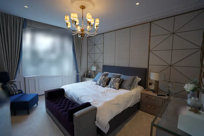 Finished bedroom with a pale textured wallcovering, geometric panelling with brass inlay behind the bed, and a crystal chandelier.