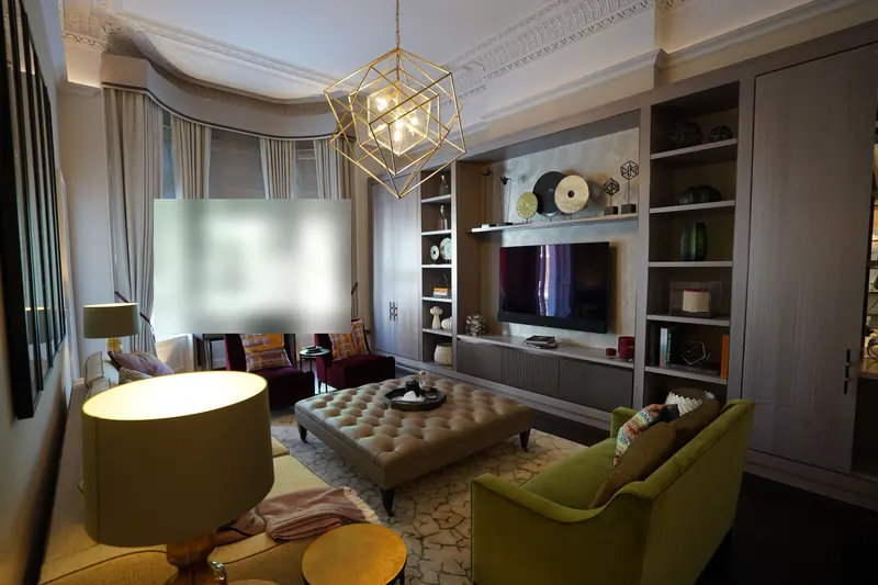 Finished drawing room with pale textured walls, a gold geometric pendant, a buttoned leather ottoman and velvet seating in green and magenta.