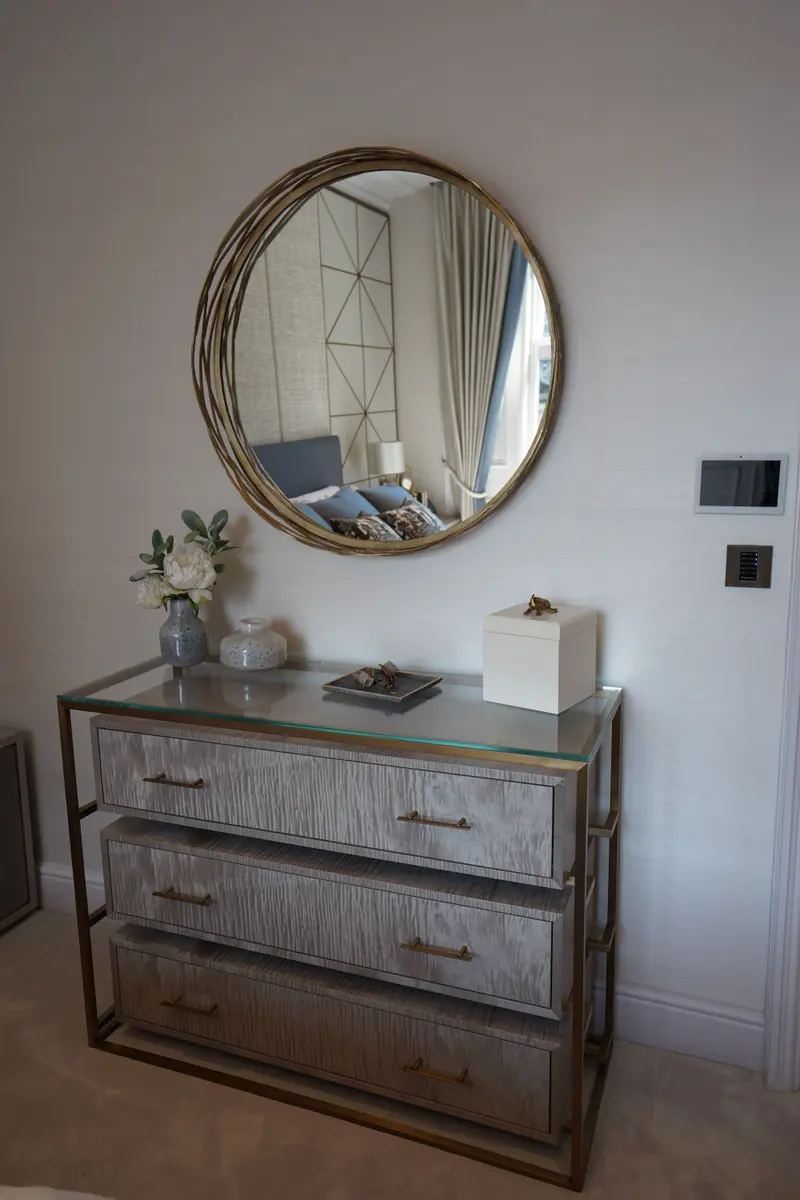 A round brass mirror above a glass-topped chest of drawers, against a pale textured wallcovering.