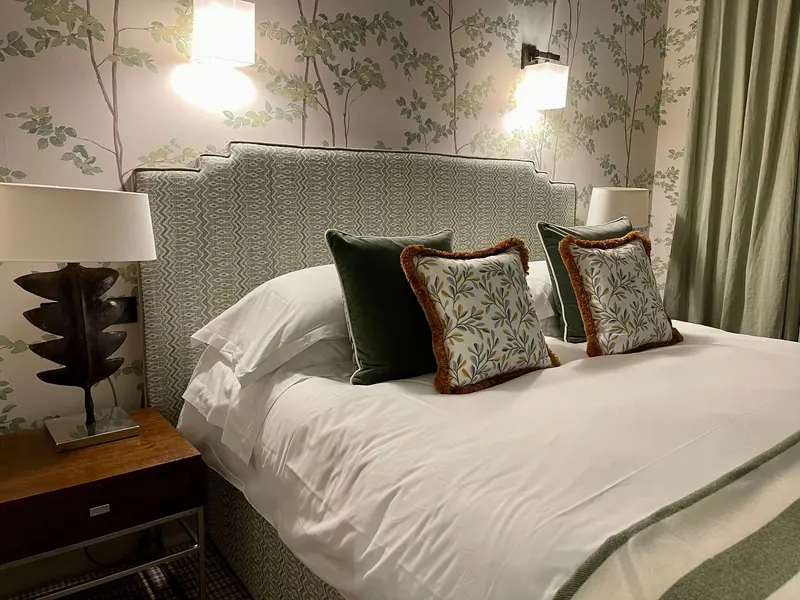 Finished guest bedroom at Brown's Hotel, Mayfair: pale tree-and-leaf wallcovering behind a green patterned headboard, with velvet and fringed botanical cushions under lit wall lights.