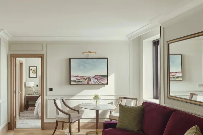 The living room of an Executive Suite at Jumeirah Carlton Tower: cream walls laid out in moulded panels, a landscape painting above a small dining table, a large wall mirror, a burgundy sofa, and a doorway through to the bedroom.