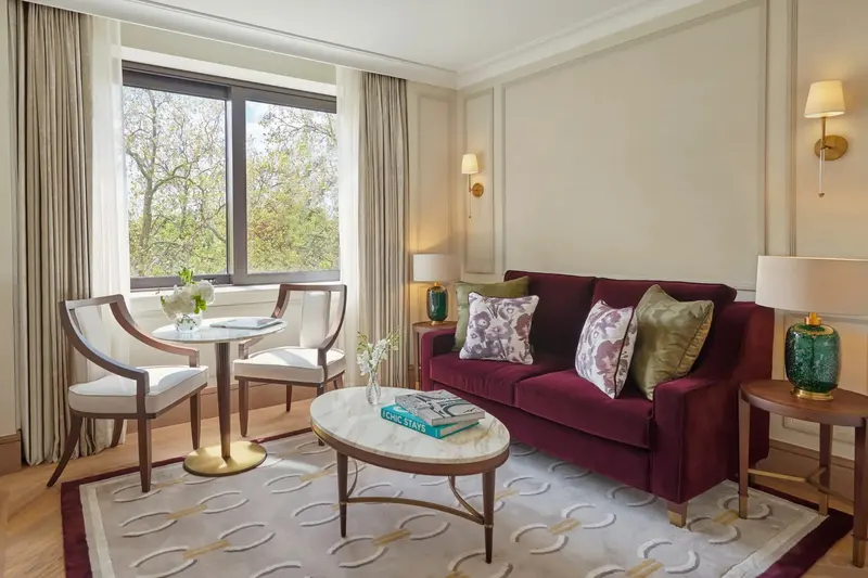 The sitting room of a Garden Suite at Jumeirah Carlton Tower: a burgundy velvet sofa with patterned cushions, an oval stone coffee table on a cream rug, brass wall lights on pale panelled walls, and a window table looking out over Cadogan Gardens.