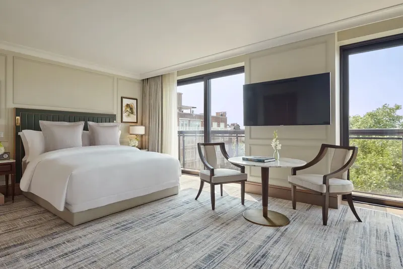 A Junior Suite at Jumeirah Carlton Tower: a large bed with a green headboard, cream wall panels framed in mouldings, a small round table and two chairs, and floor-to-ceiling windows onto a balcony.
