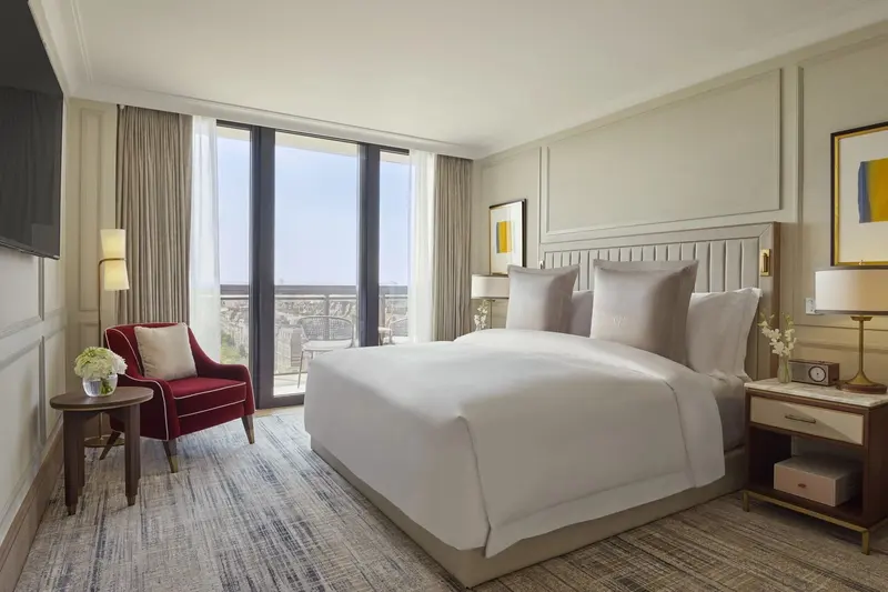 A bedroom in a Two Bedroom Luxury Suite at Jumeirah Carlton Tower: a wide bed with a pale ribbed headboard, moulded wall panels with inset wallcovering on every wall, a red armchair, and full-height windows to a balcony above the city.