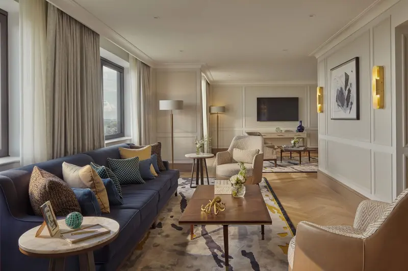 The long living room of the Royal Suite at Jumeirah Carlton Tower: a navy sofa with patterned cushions, a walnut coffee table, brass wall lights and framed art on pale panelled walls, and a dining area beyond on a herringbone oak floor.
