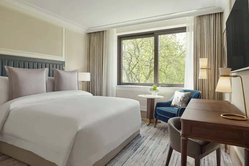 A Superior Room at Jumeirah Carlton Tower: a made-up double bed against a deep green upholstered headboard, pale panelled walls with inset wallcovering, a walnut desk with a brass lamp, and a blue armchair by a window onto the trees of Cadogan Place.