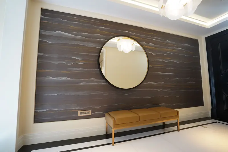 Finished entrance hall in a private residence at Ten Trinity Square: a wall of dark, banded wallcovering with streaks of silver and gold, framed in pale panelling, behind a round mirror and a tan leather bench.