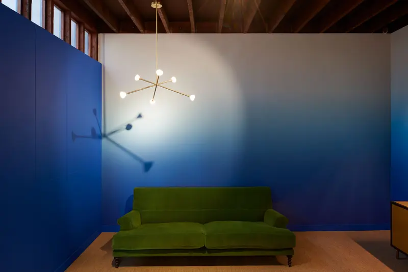 Finished showroom room: walls in a deep blue gradient mural that fades to white at the top, around a green velvet sofa under a brass pendant.