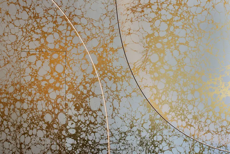 Close view of the gilded marbled wallcovering, gold veining over pale grey, crossed by the arc of a floor lamp.