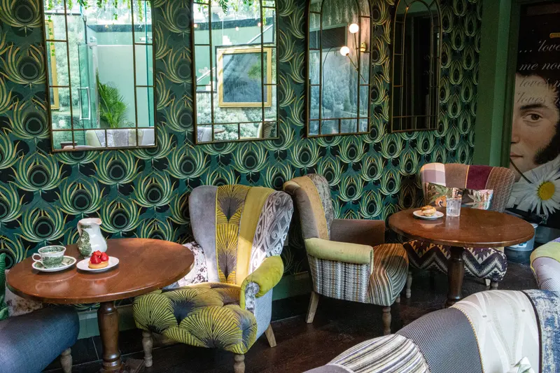 The finished tea room: a bold green-and-gold feather wallpaper on every wall, hung behind arched mirrors, patchwork armchairs and round walnut tables set for tea.