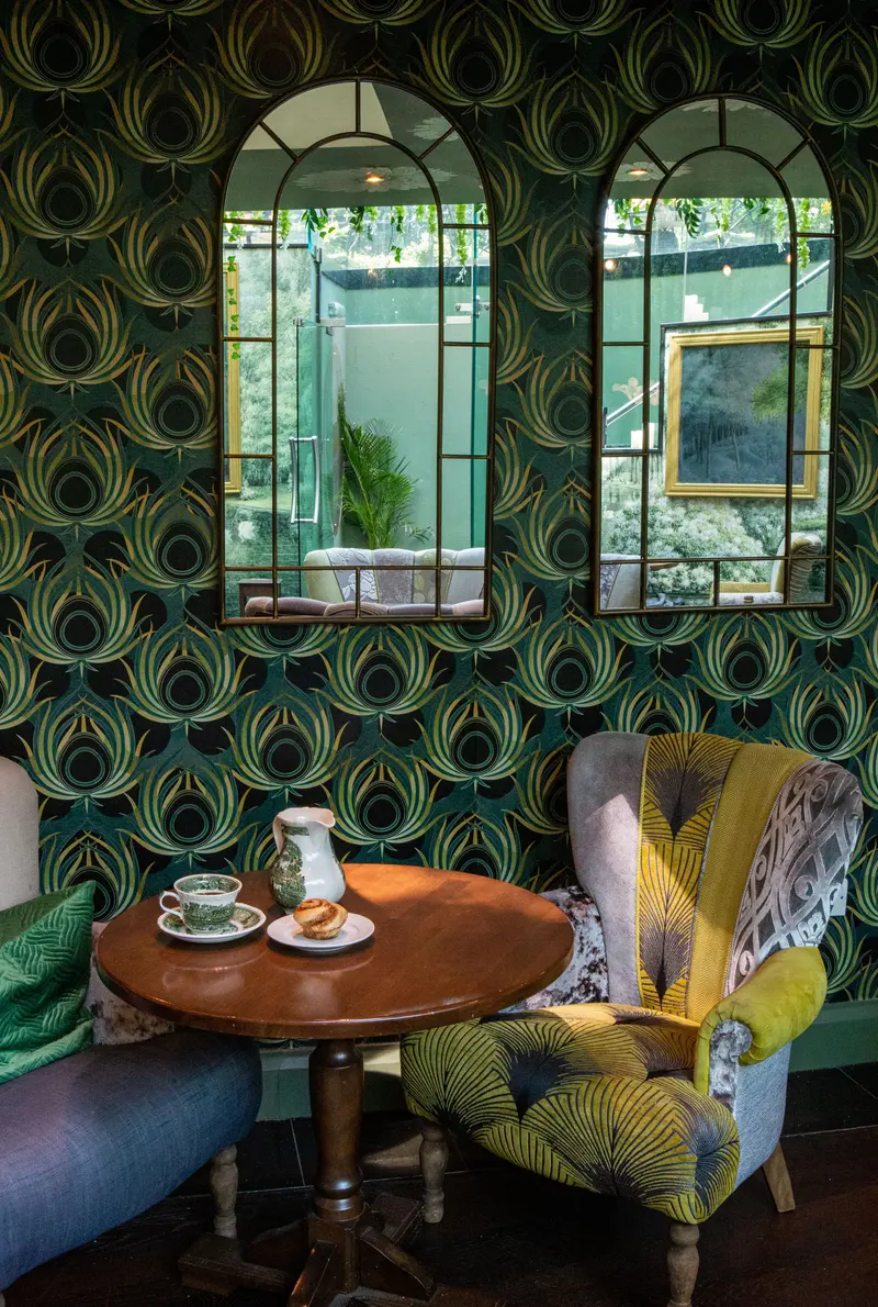Two arched mirrors set into the feather wallpaper above a table laid for tea and a feather-print armchair.
