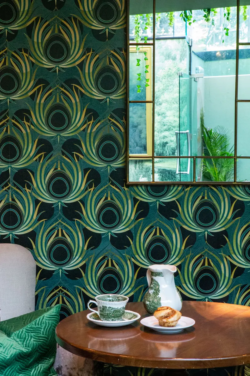Close view of the feather wallpaper beside a mirror, its gold quills crisp to the frame, above a table with a teacup and pastry.