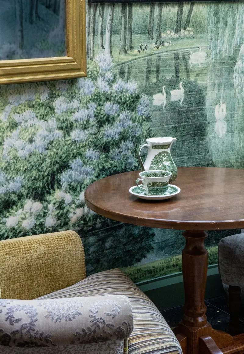 Detail of the scenic swan-lake wallpaper behind a tea table, its hydrangeas and swans running beneath a gilt frame.