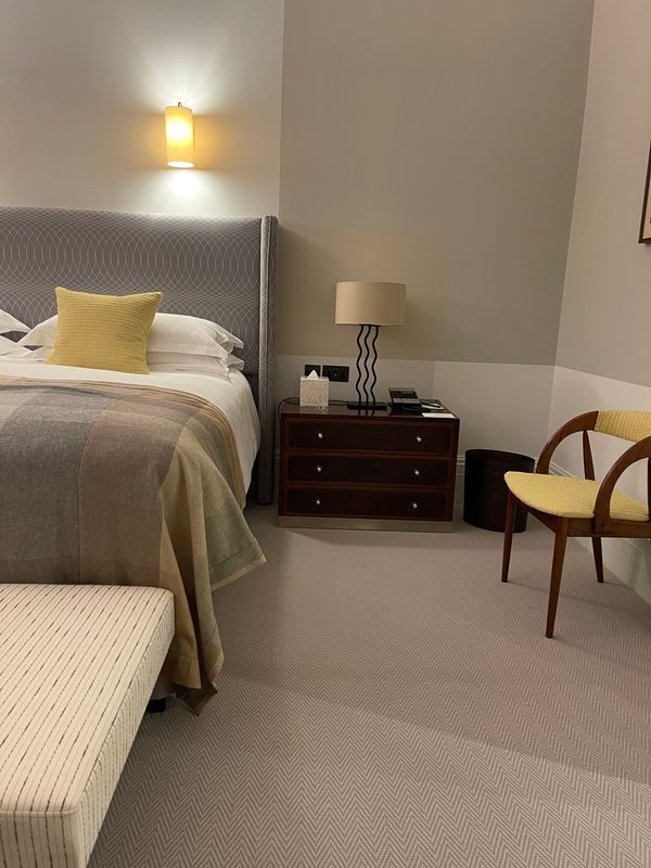 Bedside in a finished guest room at Brown's Hotel, Mayfair: a dark wood chest, a wavy-stemmed table lamp and yellow cushions against pale grey painted walls.