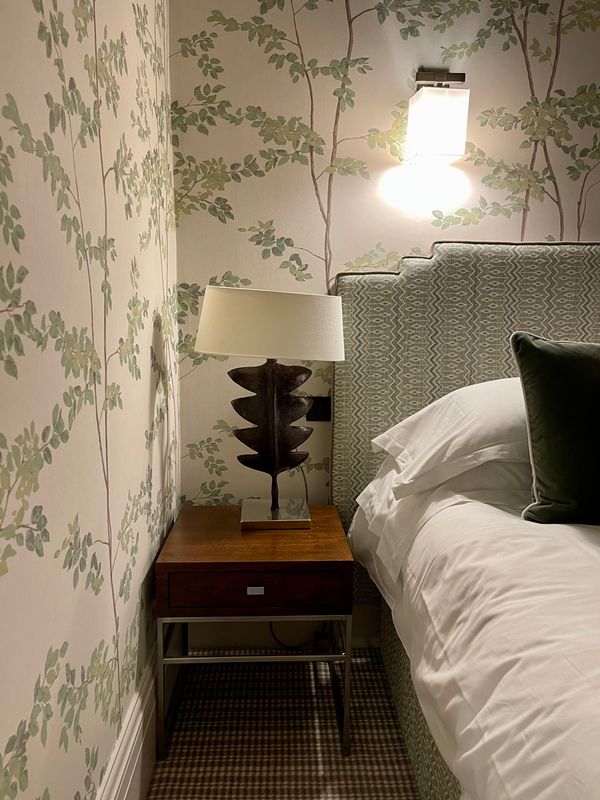Narrow bedside at Brown's Hotel, Mayfair, papered in pale tree-and-leaf wallcovering, with a sculptural bronze lamp, a wall light and a green patterned headboard.